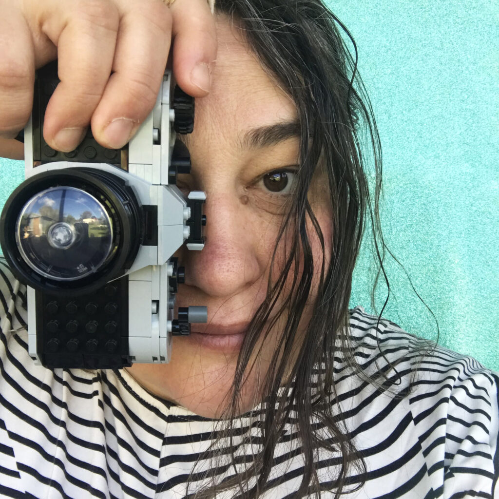 A white woman face, brown hair frames the right hand side of her face. She is holding a camera made of lego in from of the left of her face.